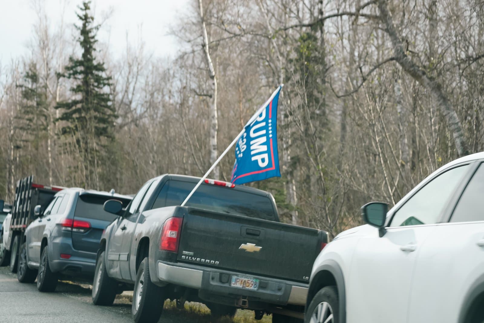 A black Chevrolet Silverado pickup truck with a Trump campaign flag attached to the bed sits in a line of vehicles.