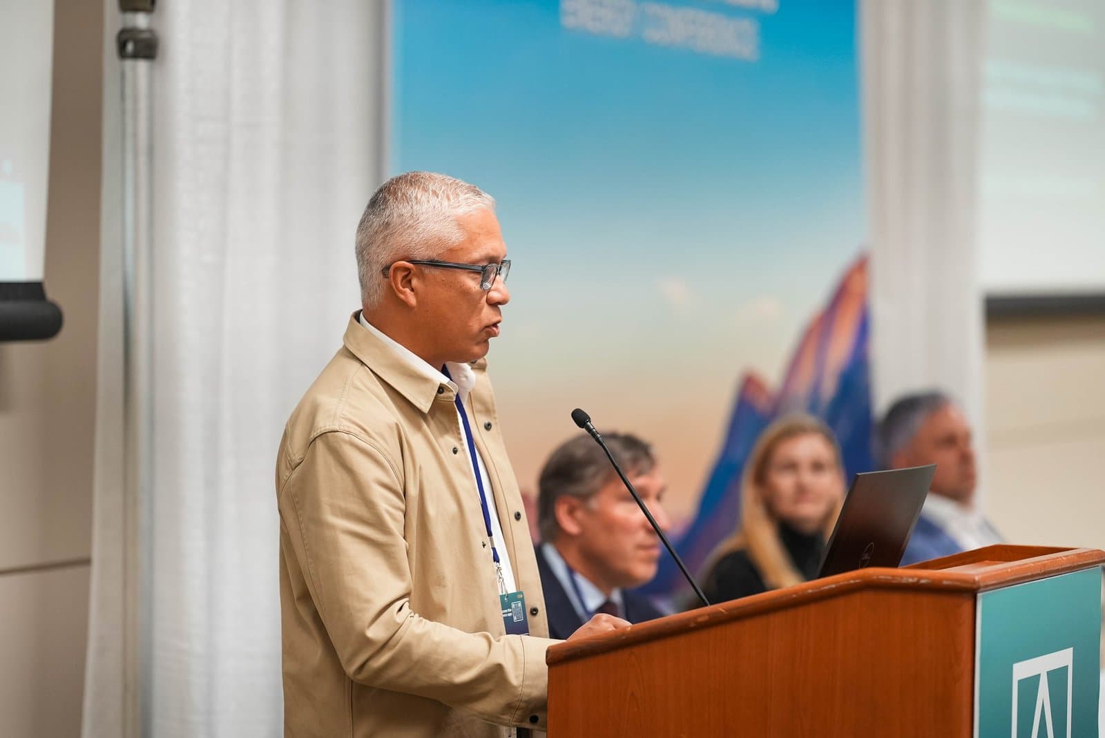 A man with gray hair and glasses, wearing a tan jacket, speaks at a wooden podium with a microphone during the Alaska Sustainable Energy Conference in Anchorage. A large banner with a blue sky and stylized mountains is visible in the background.