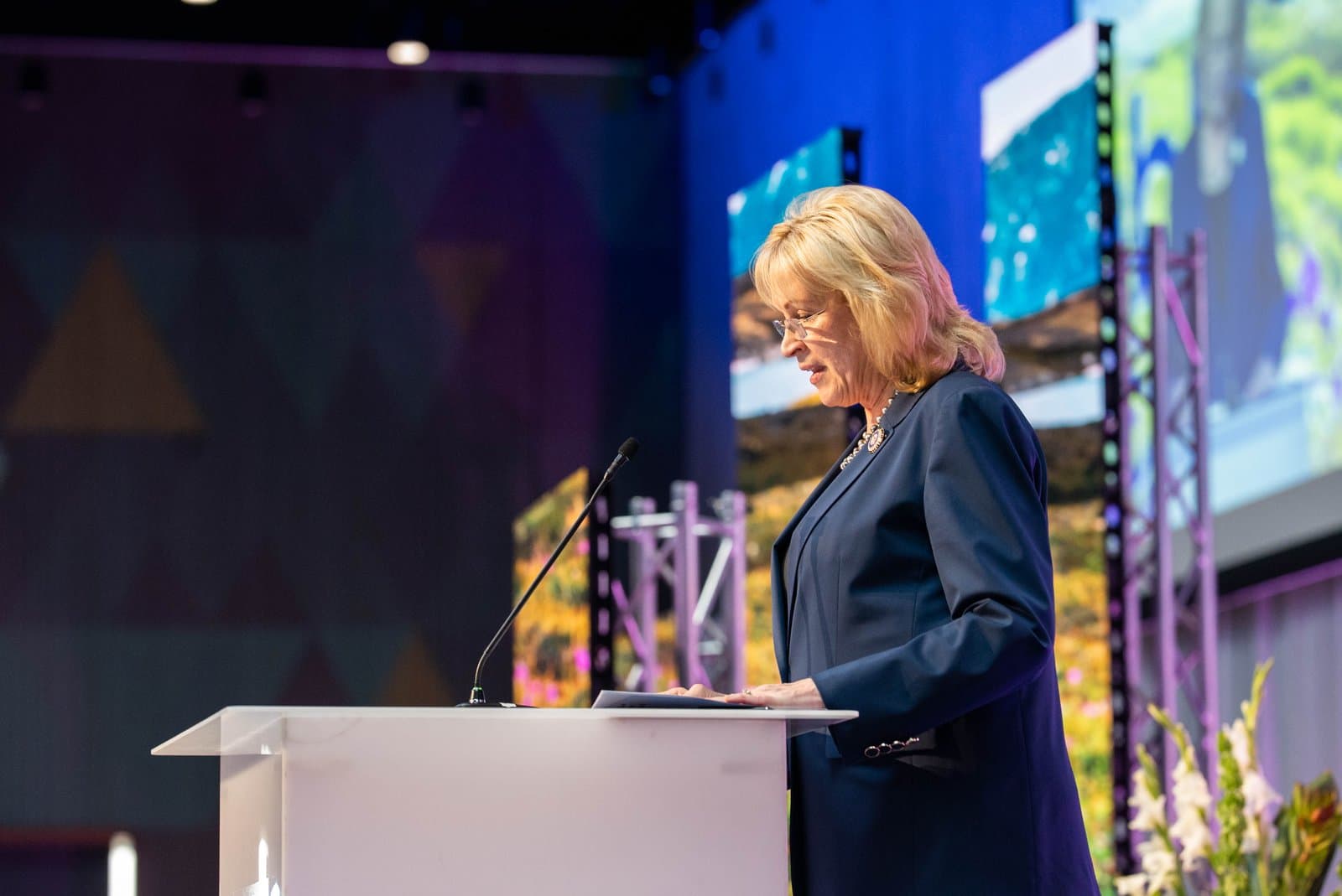 A blonde woman in glasses and a dark suit stands at a white podium, speaking into a microphone. Her hands rest on papers on the podium, and large screens displaying outdoor scenery are visible in the background.