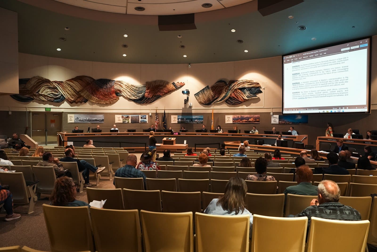 The Anchorage Assembly holds a public meeting in a circular chamber with a large projection screen displaying legislative text.