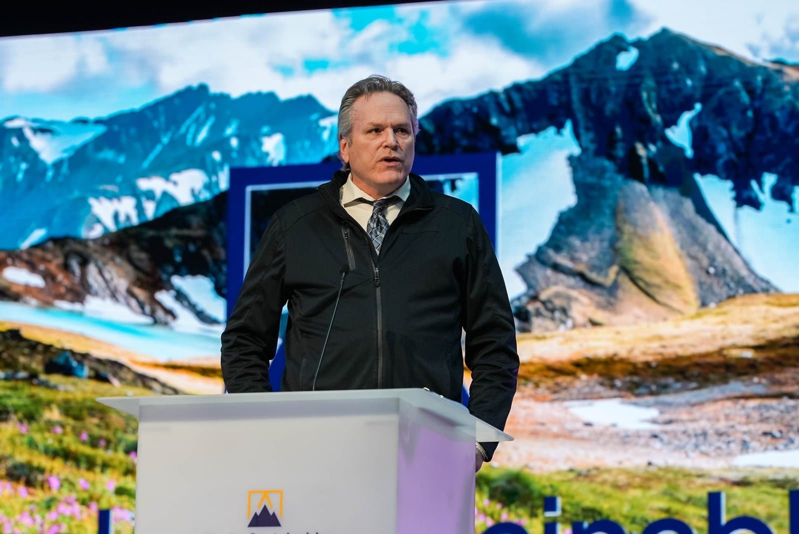 A man in a black jacket and tie speaks at a white podium during the Alaska Sustainable Energy Conference in Anchorage, with a large screen displaying a scenic mountain and glacier landscape behind him.
