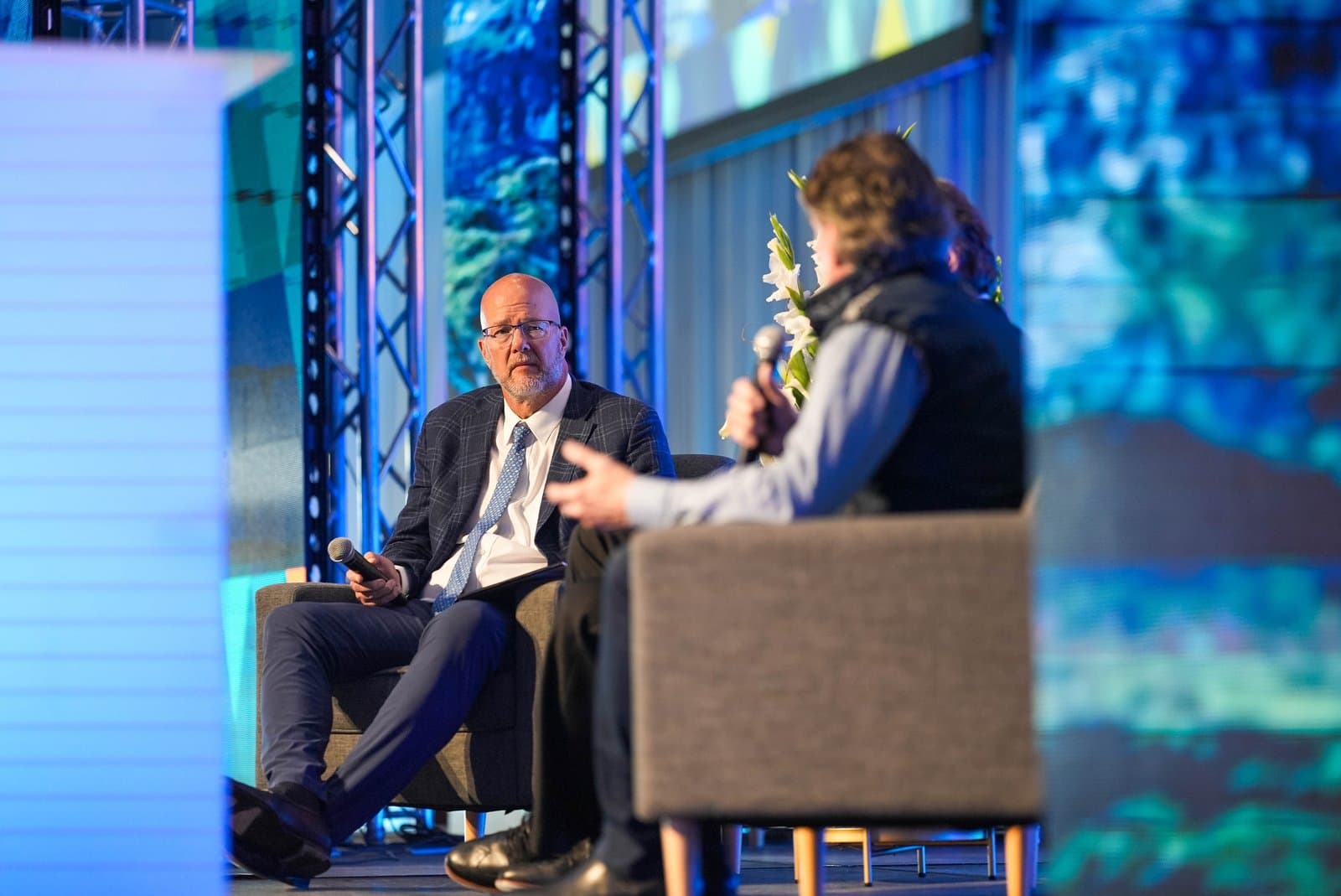 Two men are seated on a stage during a conference. The man on the left, wearing a plaid suit jacket and glasses, holds a microphone and looks towards the man on the right, who is partially visible and holding a microphone. Blue-lit screens and stage structures are in the background.