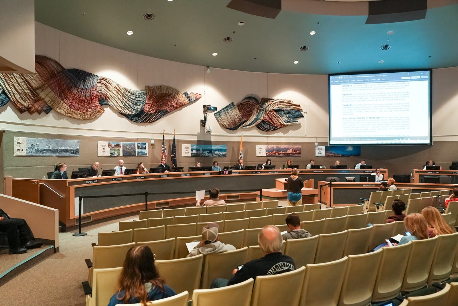 Members of the Anchorage Assembly sit at a curved dais during a public meeting held in a municipal chamber in Anchorage, Alaska.