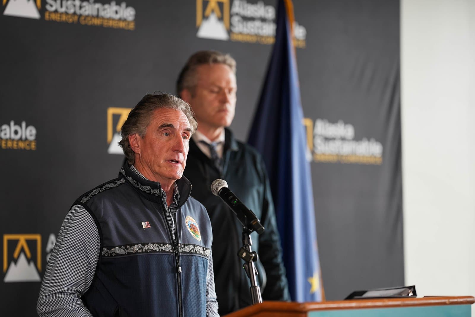 A man in a patterned vest and collared shirt speaks into a microphone at a podium, with another man and an Alaskan flag visible in the background. A banner for the 'Alaska Sustainable Energy Conference' is behind them.