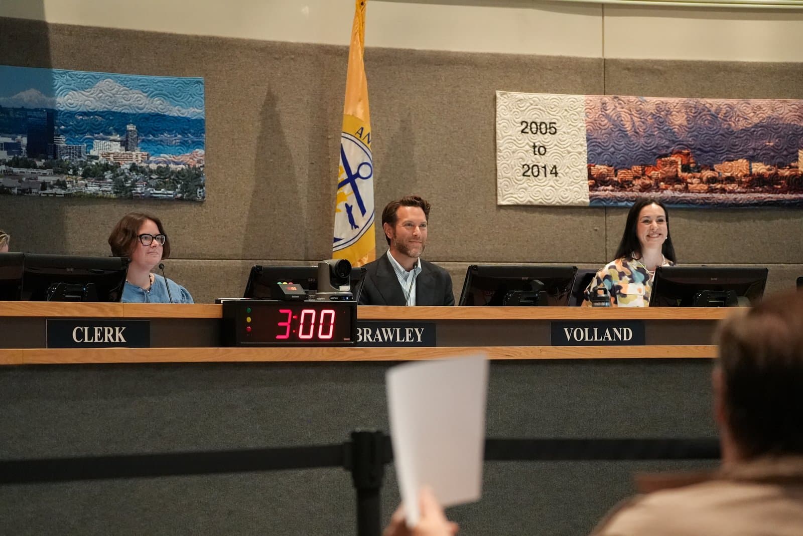 Three officials sit at a dais during an Anchorage Assembly meeting, with a digital timer displaying three minutes in the foreground.