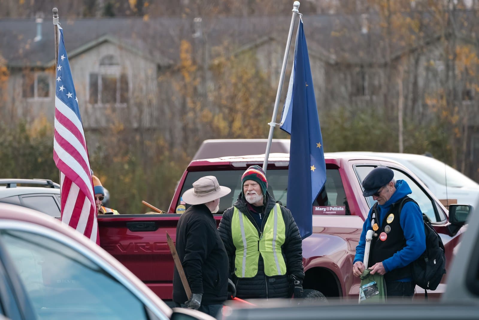 Three individuals stand near a red pickup truck displaying the American flag and the Alaska state flag during an outdoor community event.
