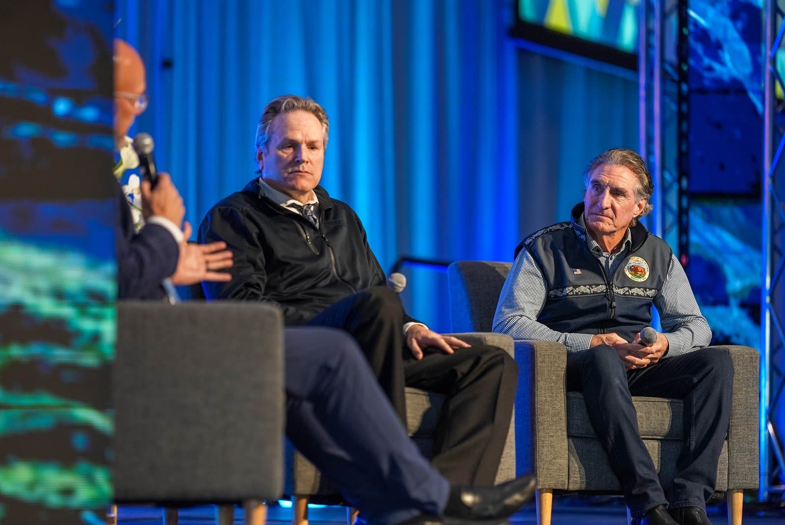 Three men sit on a stage during a panel discussion at the Alaska Sustainable Energy Conference in Anchorage. One man on the left is partially visible holding a microphone, while two other men in the center and right listen attentively, seated in grey chairs against a blue backdrop.