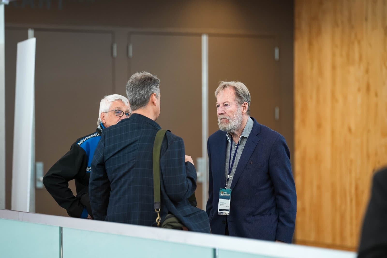 Three men are engaged in conversation at an indoor conference. The man on the right wears a blue blazer and a conference badge for the Alaska Sustainable Energy Conference, while another man with his back to the camera stands in the center.