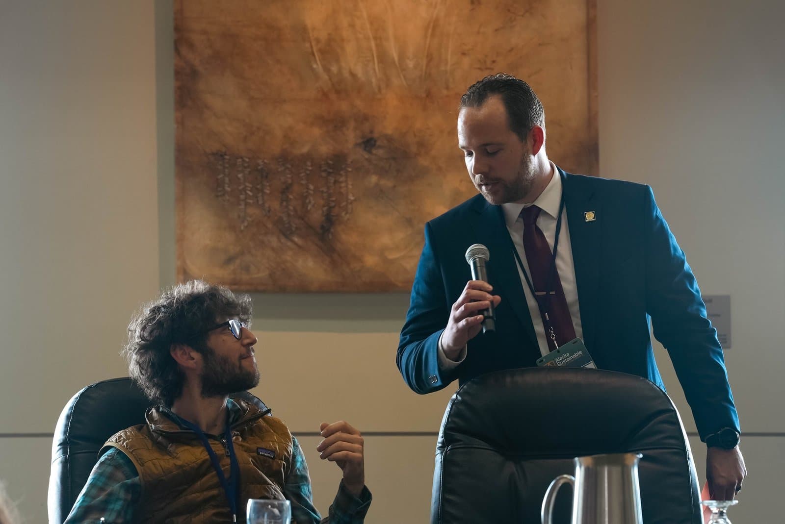 Two men participate in a discussion at an indoor conference. A man in a suit and tie holds a microphone, speaking to a seated man in glasses, a plaid shirt, and a brown vest.