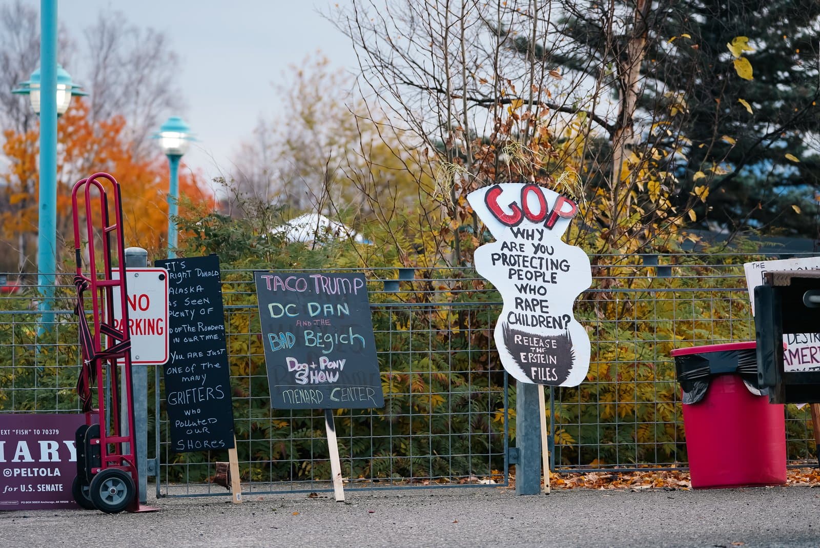 Handmade political protest signs are displayed on a chain-link fence in front of autumn foliage and a red trash bin.