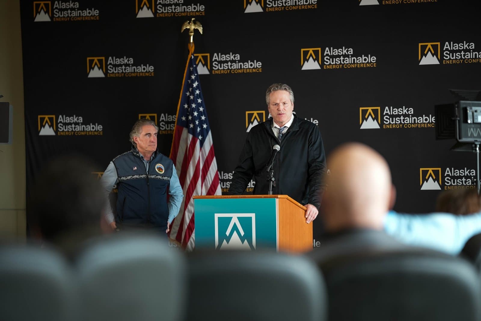 Two men stand at a podium during the Alaska Sustainable Energy Conference in Anchorage. One man, wearing a dark jacket and tie, speaks into a microphone, while another man in a blue vest stands to his left. An American flag and a backdrop with the 'Alaska Sustainable Energy Conference' logo are visible behind them.