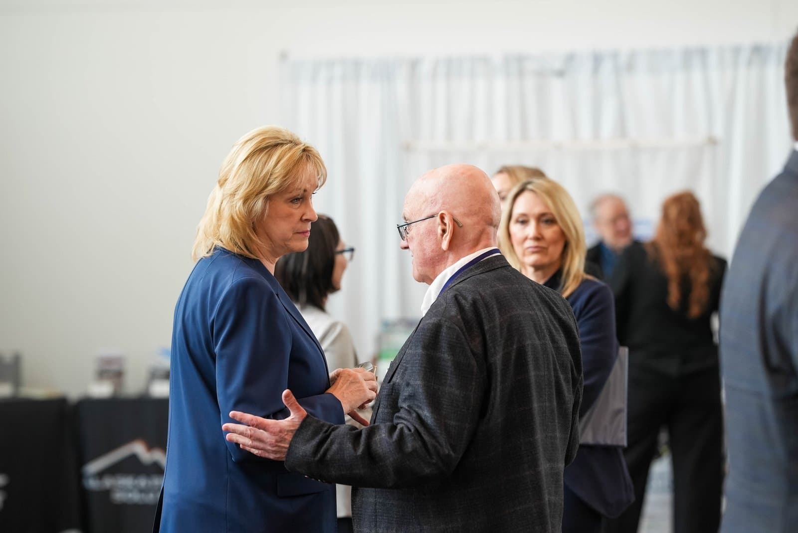 A blonde woman in a blue blazer and a bald man in a dark jacket converse at an indoor conference, with other attendees blurred in the background. A banner with a mountain logo is partially visible on the left.