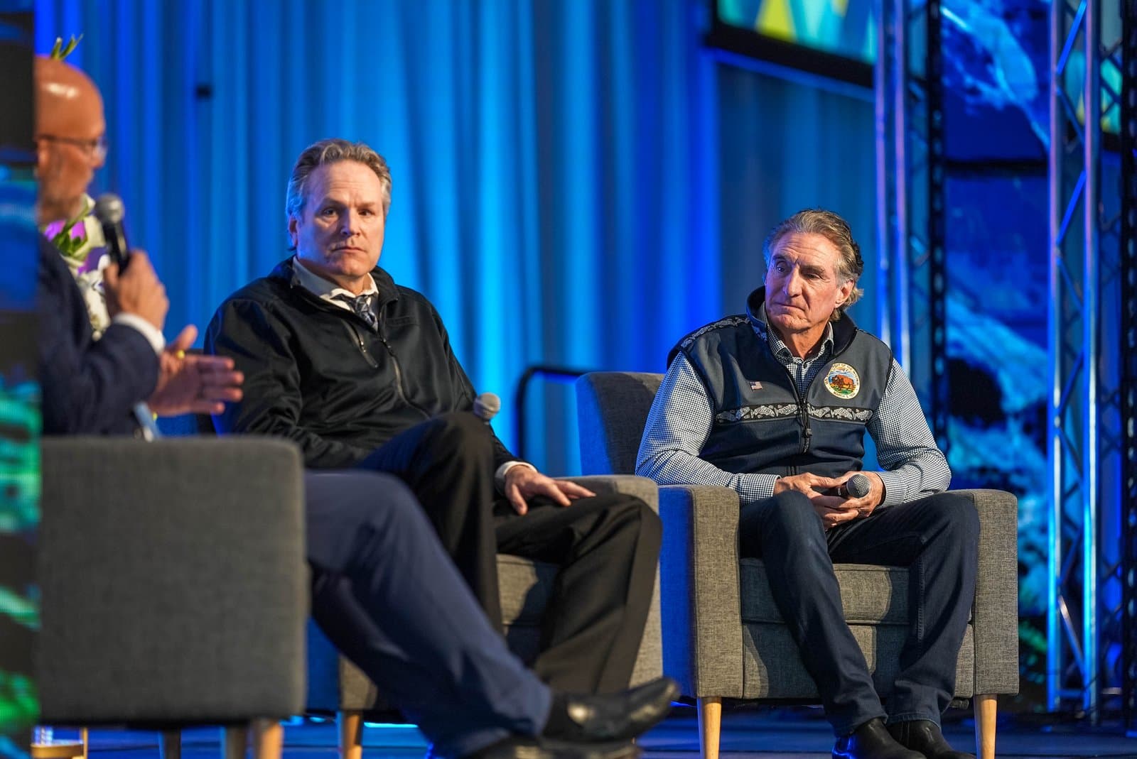 Three men, including two seated panelists and one partially visible moderator holding a microphone, engage in a discussion on a stage with a blue backdrop at the Alaska Sustainable Energy Conference in Anchorage.