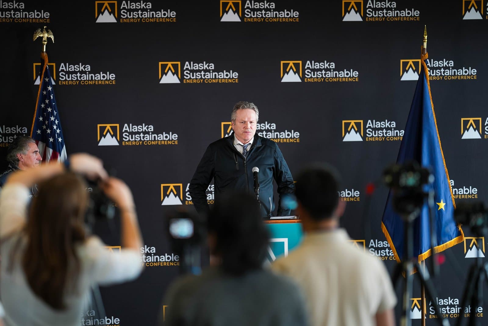 Alaska Governor Mike Dunleavy speaks at a podium during the Alaska Sustainable Energy Conference, with American and Alaskan flags on either side and a branded backdrop.