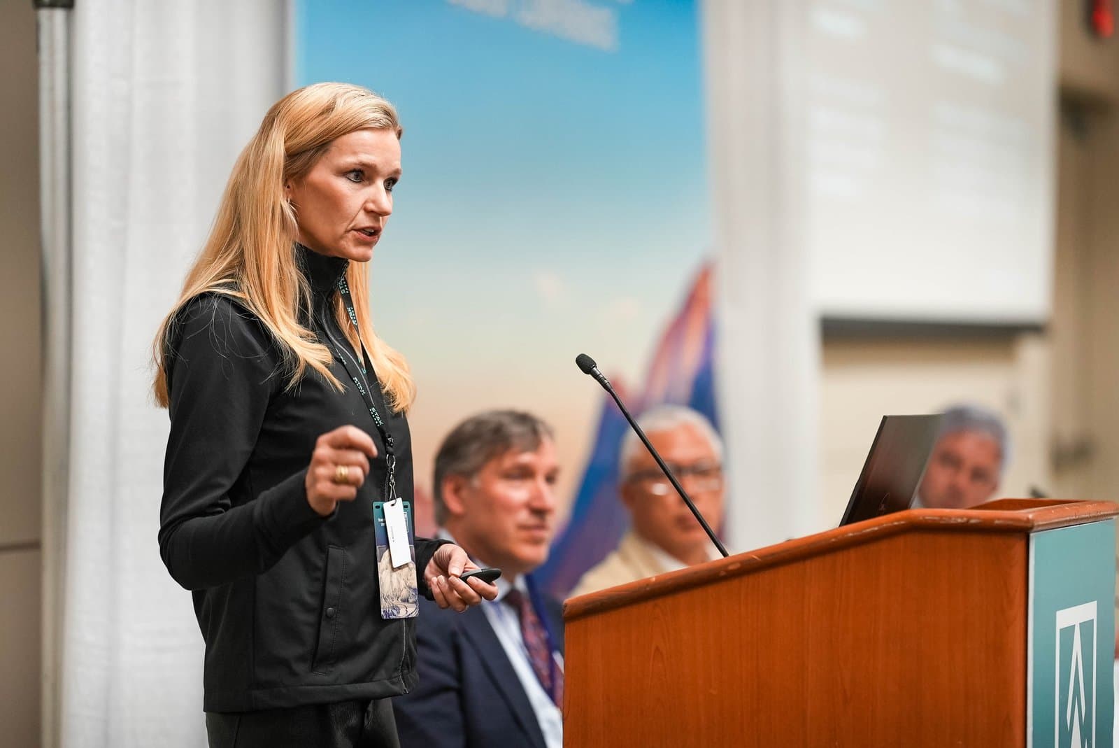 A woman with long blonde hair, wearing a black top and a lanyard, speaks at a wooden podium with a microphone. She holds a remote control, addressing an audience at the Alaska Sustainable Energy Conference in Anchorage.
