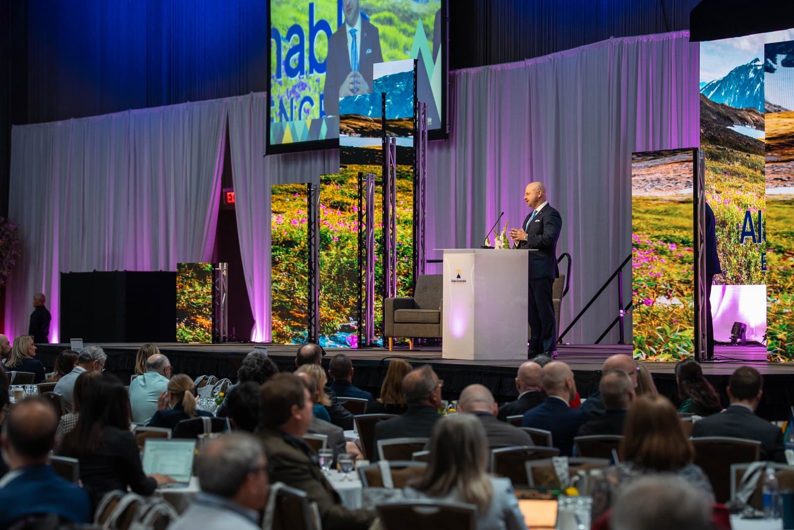 A speaker addresses an audience at the Alaska Sustainable Energy Conference in Anchorage, with stage screens displaying Alaskan landscapes and a main screen showing a presenter.