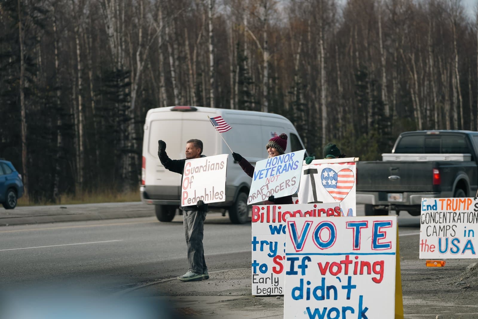 Protesters stand on a roadside holding political signs with messages about the GOP and voting while a white van drives past in the background.