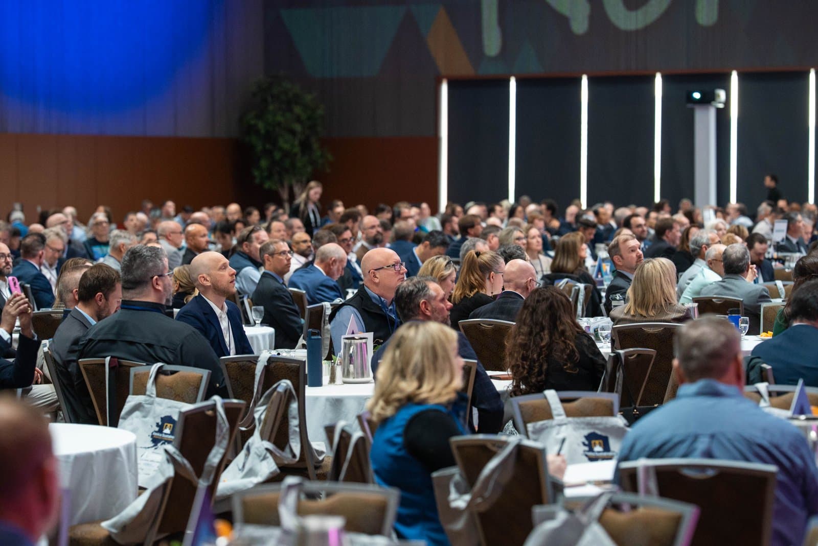 A wide shot of a large indoor conference room filled with many attendees seated at round tables, listening to a presentation. The room features modern lighting and a large screen at the front.