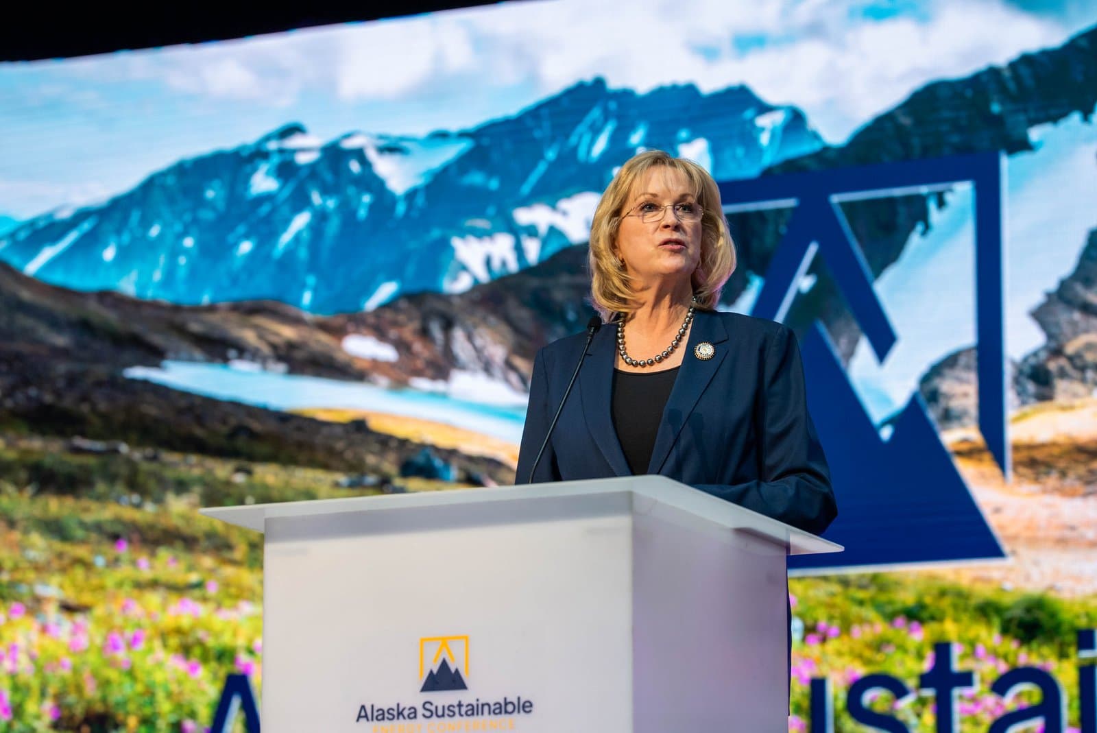 A woman with blonde hair and glasses, wearing a dark suit and pearl necklace, speaks at a podium. Behind her, a large screen displays a scenic Alaskan mountain landscape and the logo for the Alaska Sustainable Energy Conference.