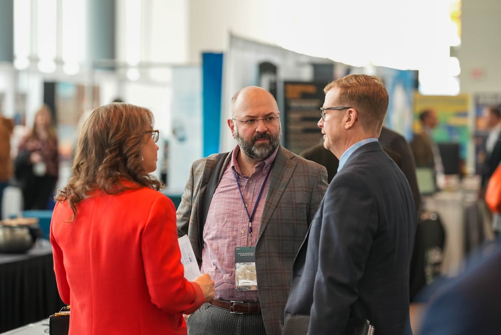 Three professionals, two men and one woman, are engaged in conversation at the Alaska Sustainable Energy Conference in Anchorage. The woman, wearing a red jacket, faces away from the camera, while the two men, one with a beard and glasses in a plaid blazer and the other in a dark suit and glasses, face each other in discussion. A conference badge is visible on one man, and other blurred attendees are in the background.