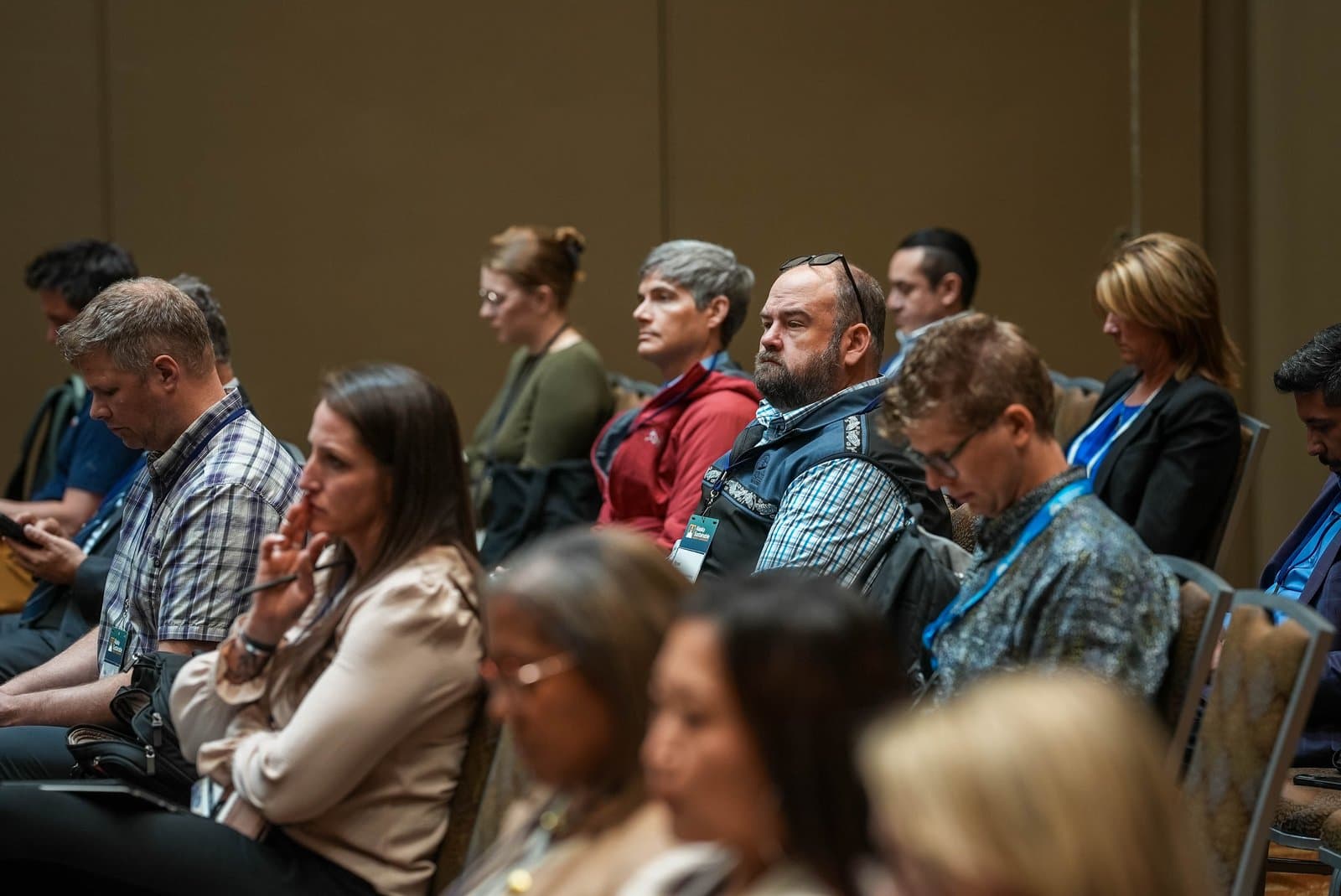 A diverse group of adults are seated in rows within a conference room, attentively listening to a presentation. Some individuals are looking forward, while others are engaged with notes or electronic devices. The room features a plain, warm-toned wall in the background.