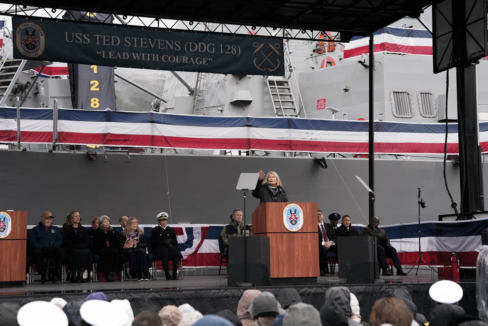 A woman speaks at a podium in front of the USS Ted Stevens (DDG 128) during a commissioning ceremony with attendees seated nearby.