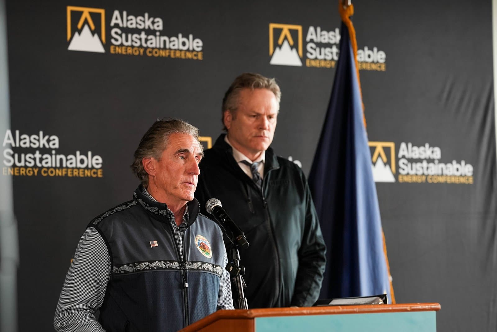 Two men stand at a podium in front of a black backdrop featuring the 'Alaska Sustainable Energy Conference' logo. An Alaska flag is partially visible on the right.