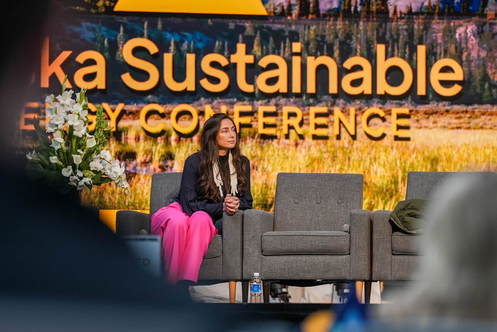 A woman with long dark hair and bright pink pants sits on a grey armchair on a stage, looking intently to her left. Behind her, a large screen displays the words "Alaska Sustainable Energy Conference" over a blurred image of a natural landscape. A bouquet of white flowers is visible on the left side of the stage.