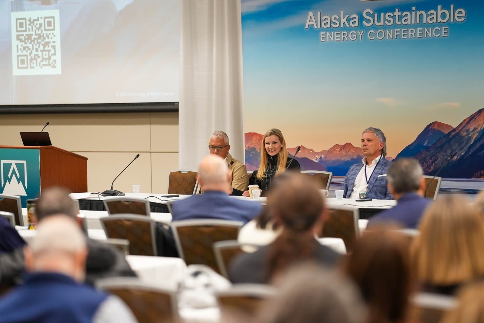 A panel of three speakers, two men and one woman, are seated at a table on a stage, addressing an audience at the Alaska Sustainable Energy Conference in Anchorage. A large backdrop behind them features a scenic Alaskan mountain and lake landscape, along with the conference title.