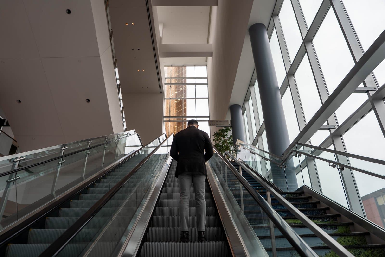 A person in a dark jacket and grey pants rides an escalator upwards inside a modern building with large windows, likely at the Alaska Sustainable Energy Conference in Anchorage.