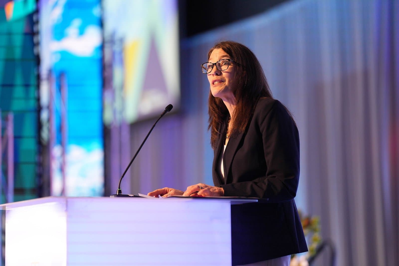 A woman with long brown hair and glasses, wearing a dark blazer over a white top, speaks at a white podium with a microphone. Her hands rest on papers on the podium, and a blurred background of blue and purple lights suggests an indoor conference setting.