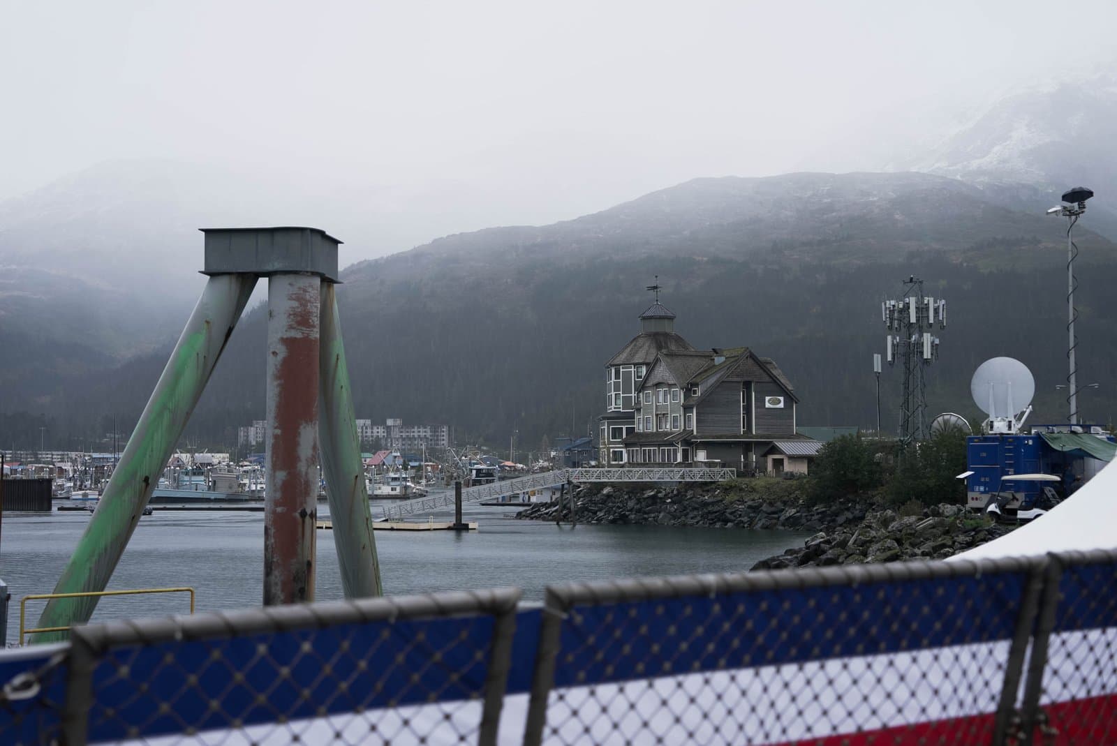 A multi-story wooden building stands on a rocky shoreline near a harbor filled with fishing vessels under a cloudy, overcast sky.