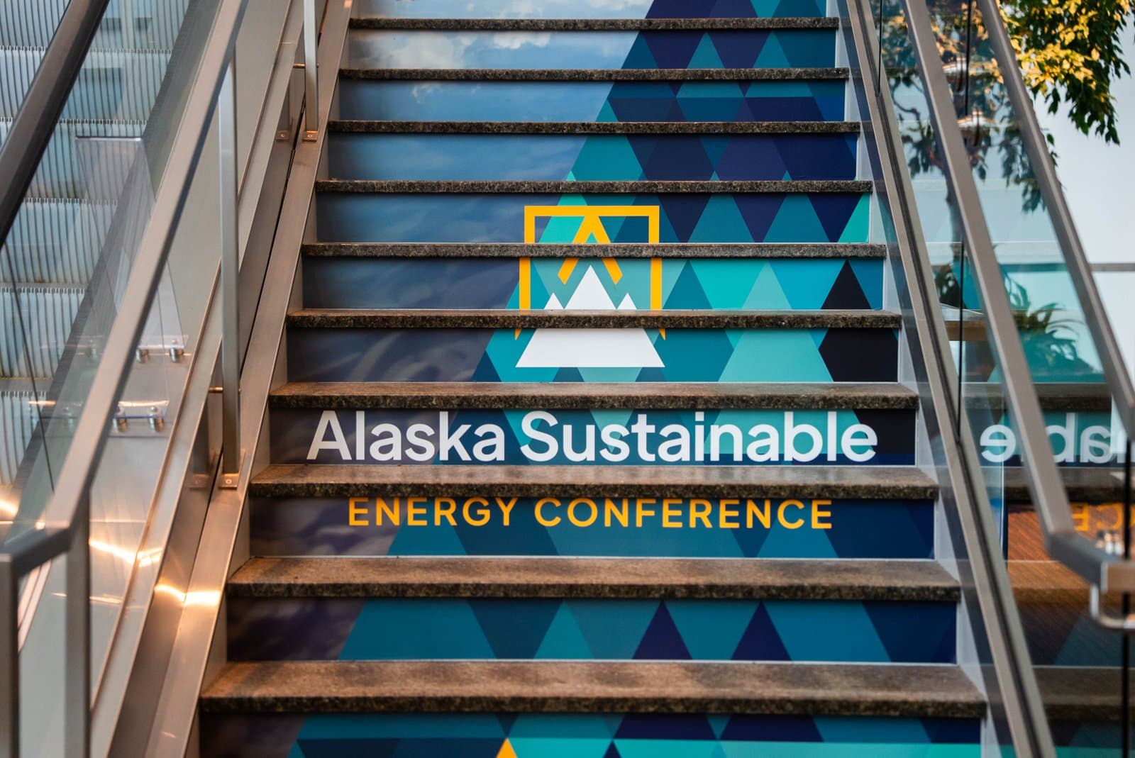 A low-angle view of a staircase featuring a large graphic display on the risers. The graphic has a blue and teal geometric pattern, a white mountain logo with an orange outline, and the text "Alaska Sustainable ENERGY CONFERENCE" in white and orange.