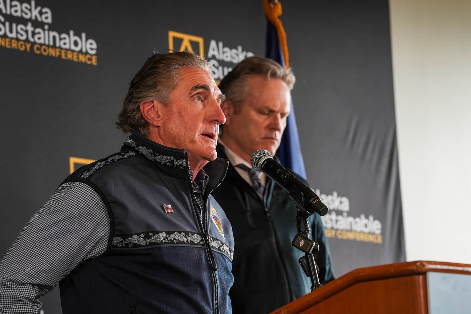 Two men stand at a podium with a microphone, one speaking, against a backdrop for the Alaska Sustainable Energy Conference.
