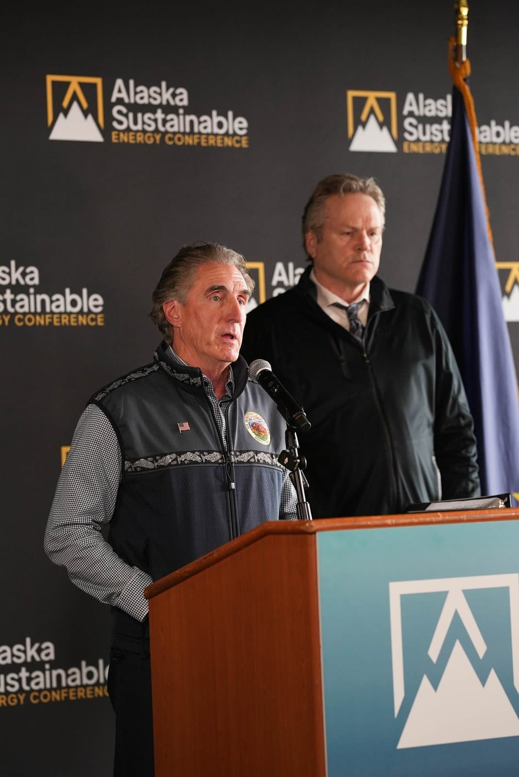 Two men stand at a podium with a microphone, speaking at the Alaska Sustainable Energy Conference. The man in the foreground wears a dark vest over a checkered shirt, with an American flag pin and a circular patch. A second man stands behind him, and a backdrop with the conference logo and name is visible, along with a partial view of the Alaska state flag.
