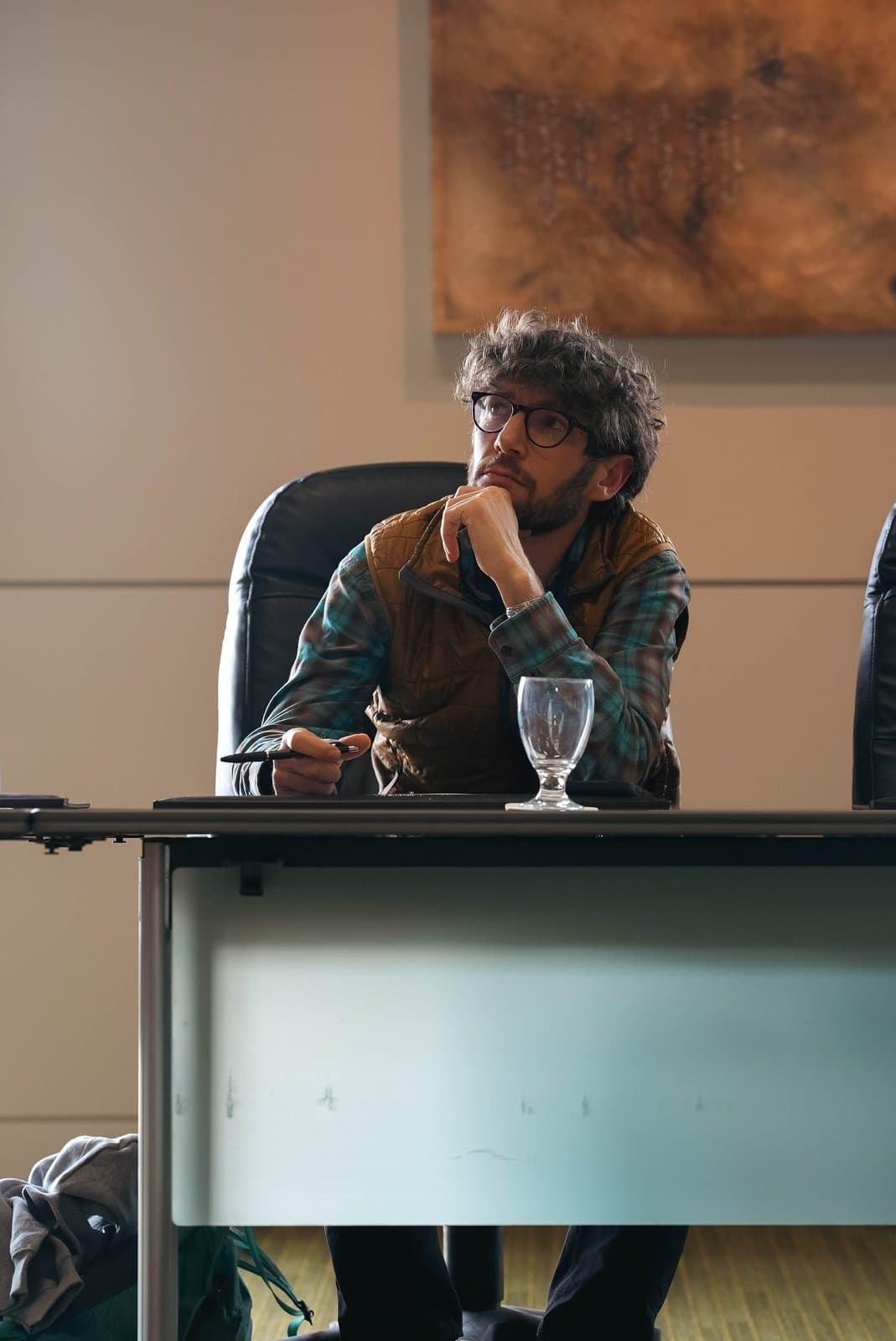 A man with curly hair and glasses, dressed in a plaid shirt and vest, sits at a conference table in Anchorage, looking thoughtfully upwards. He holds a pen, with an empty glass on the table, during the Alaska Sustainable Energy Conference.