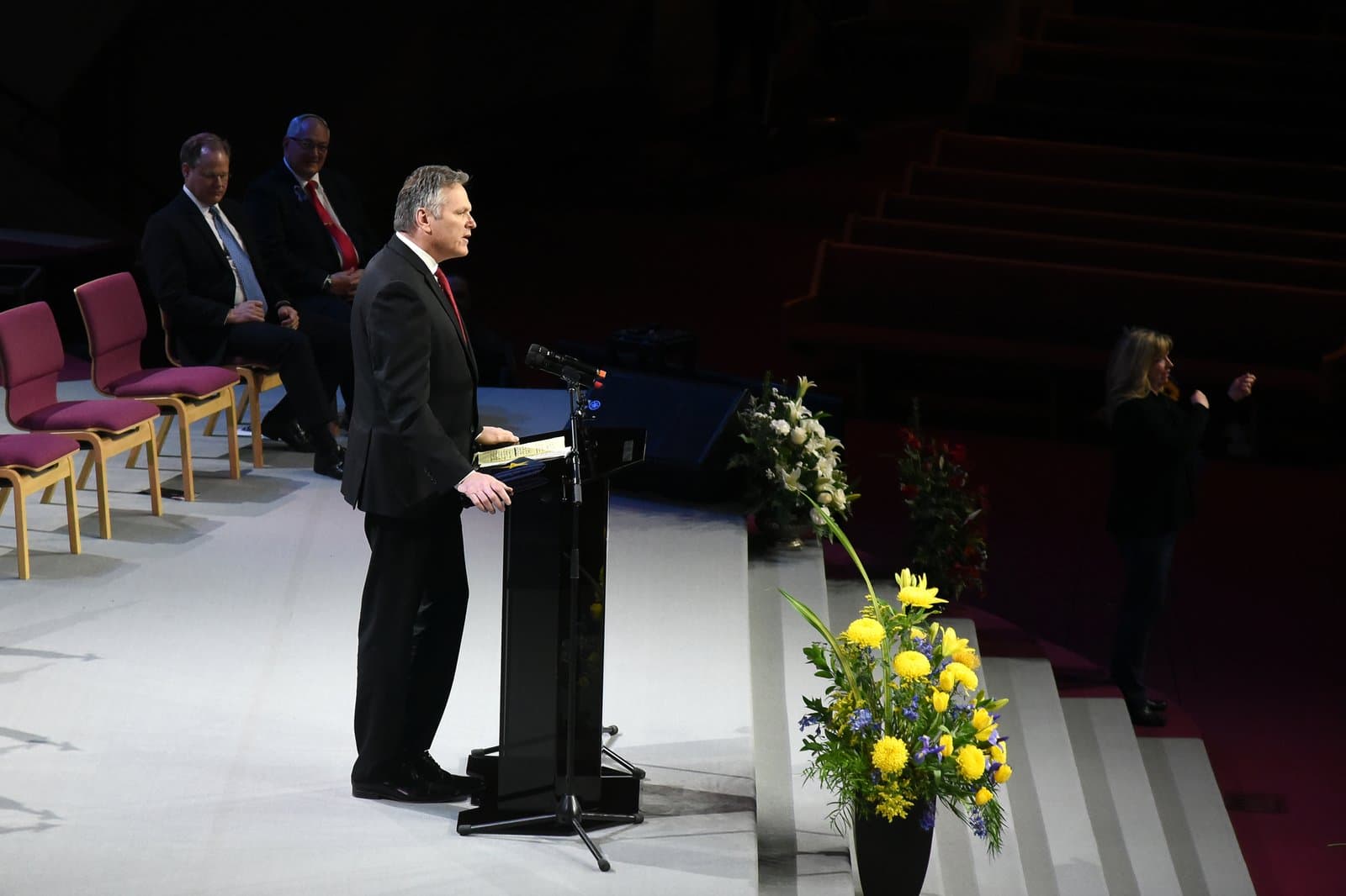 A man in a suit speaks from a podium during the memorial service for Don Young in an auditorium setting.