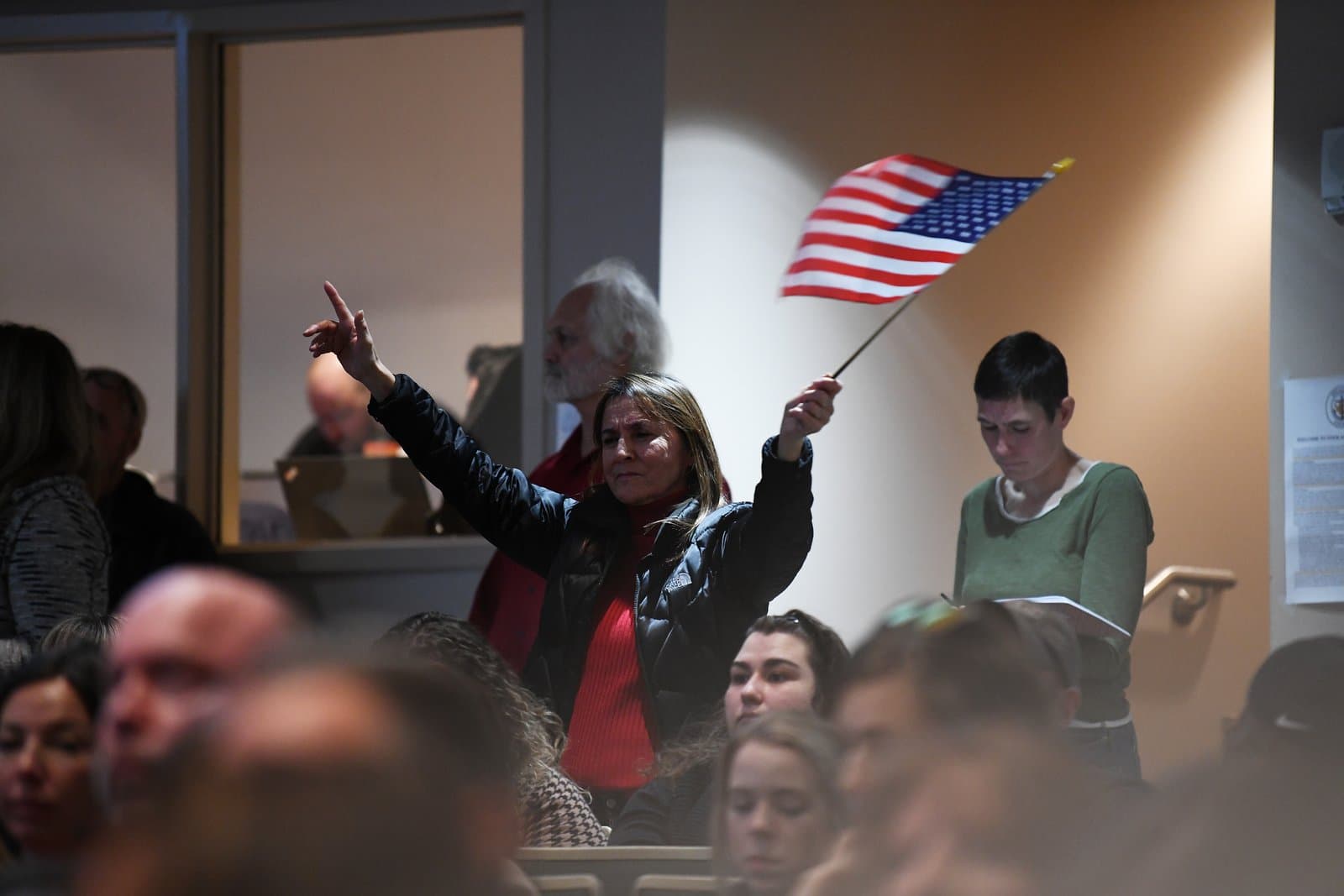A woman holds a small American flag and raises her hand during an Anchorage Assembly meeting, surrounded by other attendees in an indoor setting.