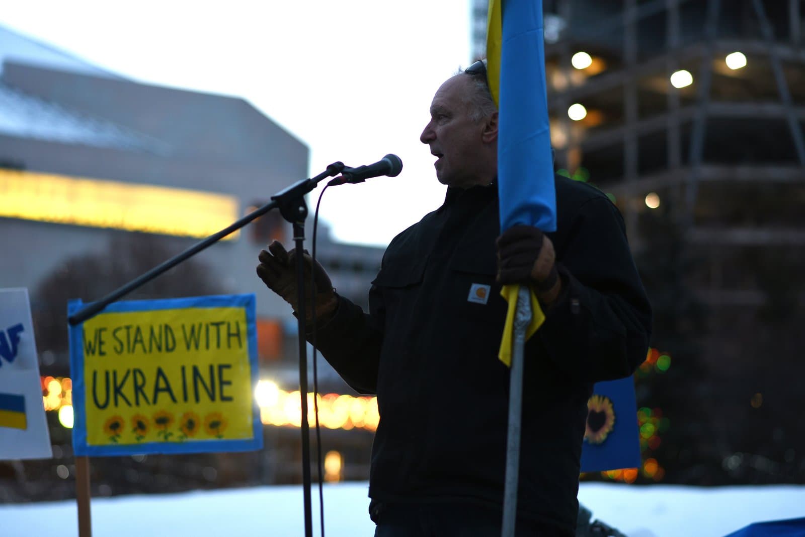 A man speaks into a microphone while holding a Ukrainian flag at an outdoor rally in Anchorage, Alaska, supporting Ukraine.