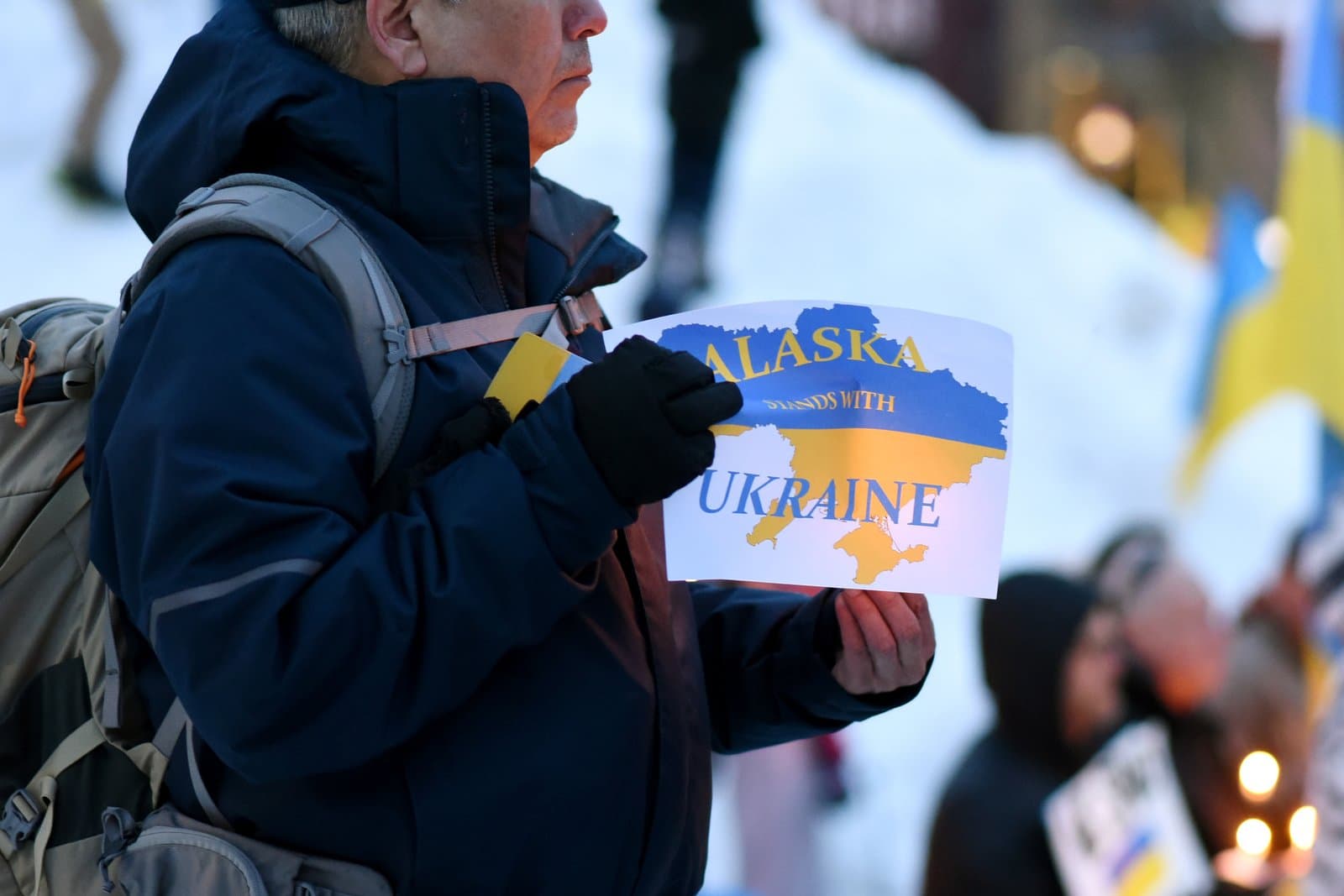 A person wearing a winter jacket and backpack holds a sign reading Alaska Stands with Ukraine during a community gathering in Anchorage.