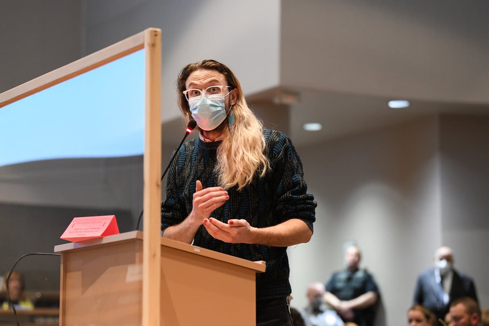 A person with long blonde hair, glasses, and a light blue face mask speaks at a wooden podium with a microphone. They are wearing a dark patterned sweater and have red nail polish on their hands, which are gesturing. A clear barrier is visible to the left, and other blurred individuals are in the background.