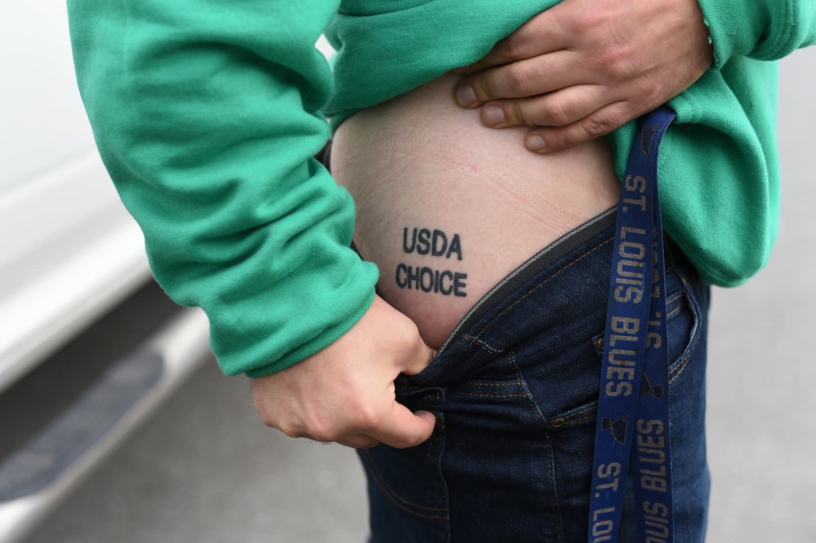 A person displays a USDA Choice tattoo on their hip while wearing a green sweatshirt and jeans with a St. Louis Blues lanyard.