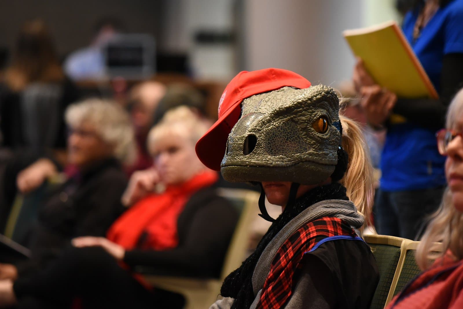 A person wearing a dinosaur mask and a red baseball cap sits in an audience at the Anchorage Assembly Meeting, with other attendees blurred in the background.