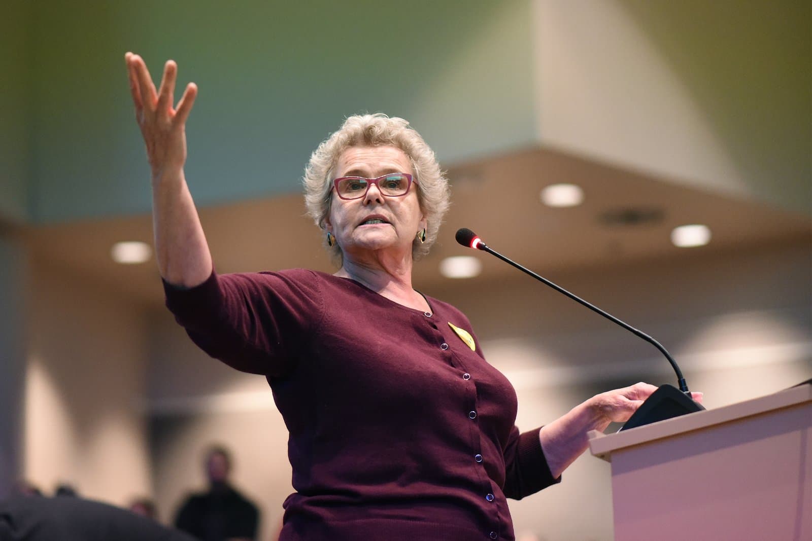 A woman with short blonde hair and red-rimmed glasses gestures while speaking at a podium during an Anchorage Assembly meeting in October 2021.