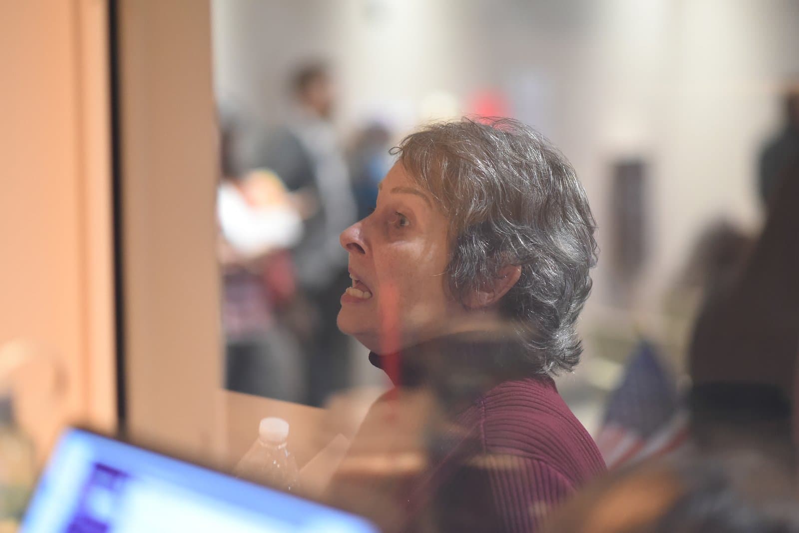 A woman with gray, curly hair, wearing a dark red turtleneck, is seen in profile, speaking or reacting during an Anchorage Assembly Meeting. The image is captured through a reflective surface, with blurred reflections of a laptop and water bottle in the foreground, and other attendees and an American flag in the background.