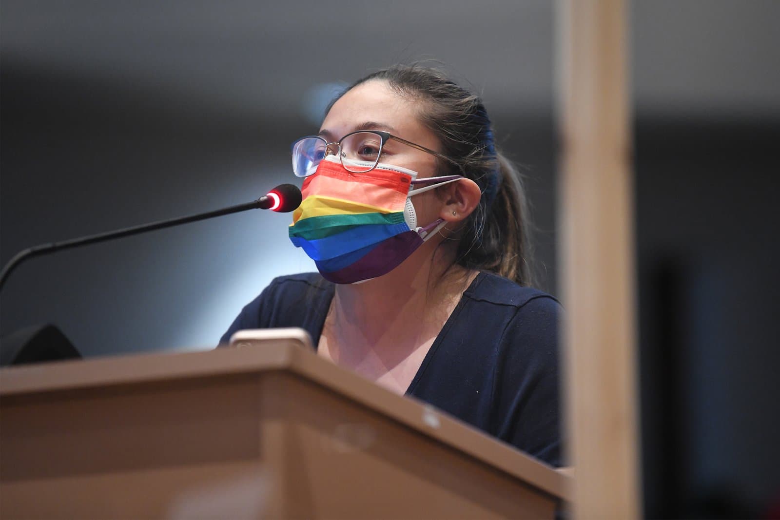 A person wearing a rainbow-patterned face mask speaks into a microphone at an Anchorage Assembly meeting in Anchorage, Alaska.