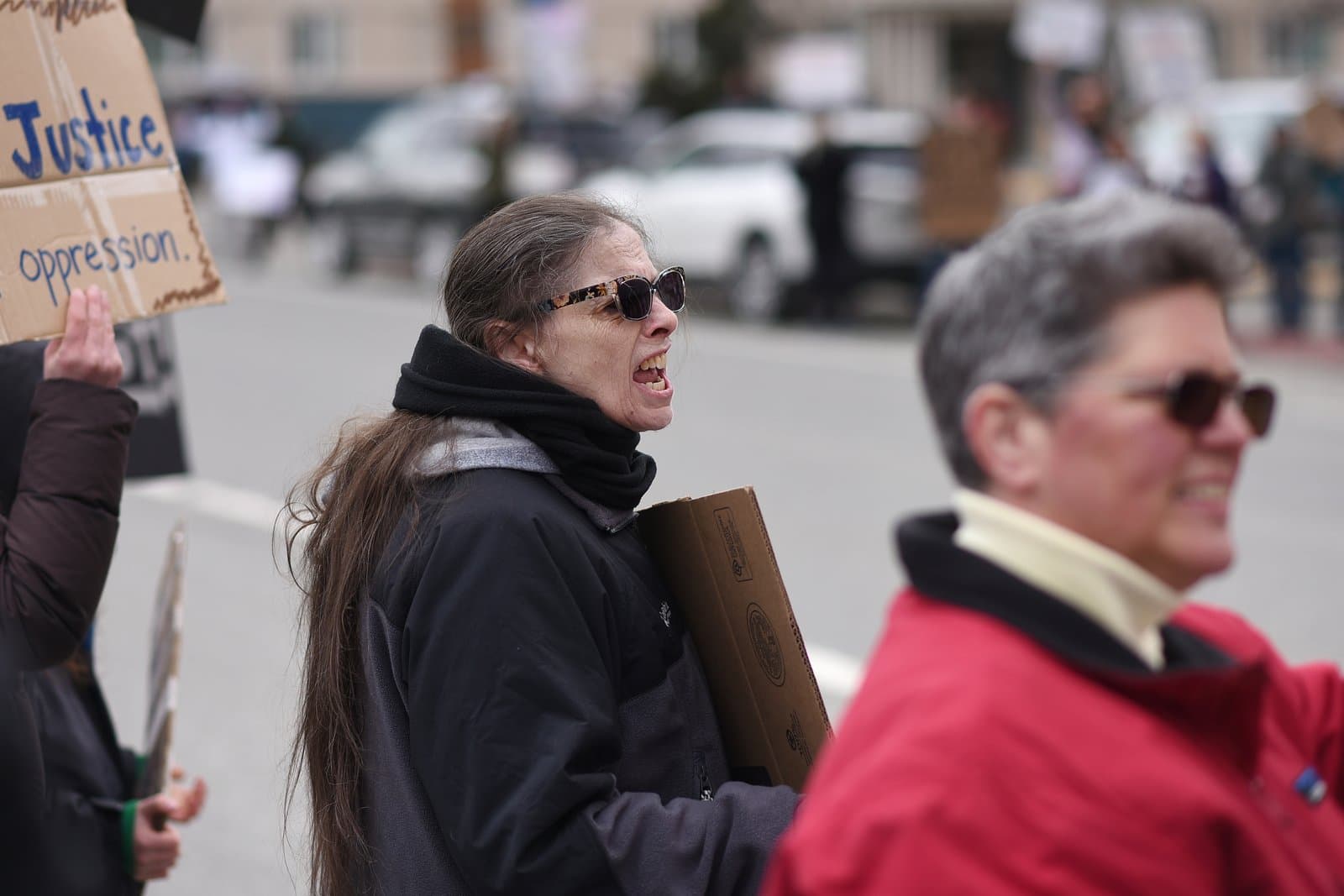 A woman with long hair and sunglasses shouts while holding a cardboard sign during a 50501 protest in Anchorage, Alaska.