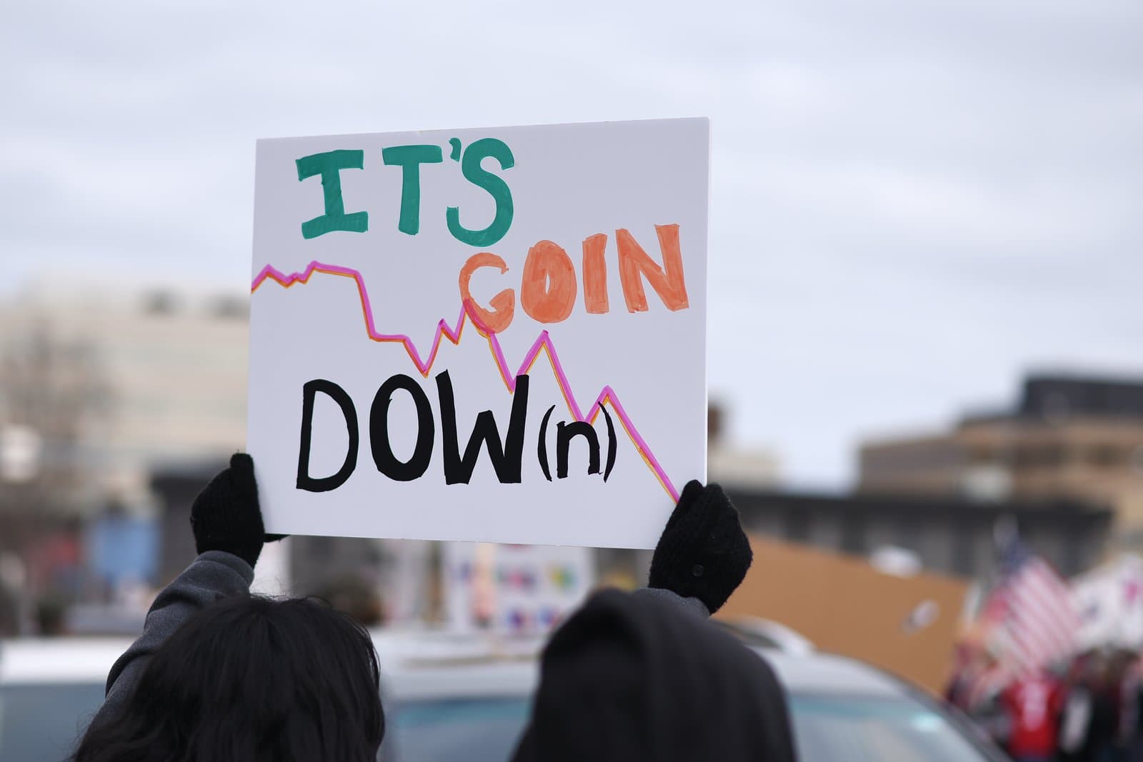 A protester holds a handmade sign reading It's Goin Dow(n) with a downward-trending line graph during a public demonstration in Anchorage, Alaska.