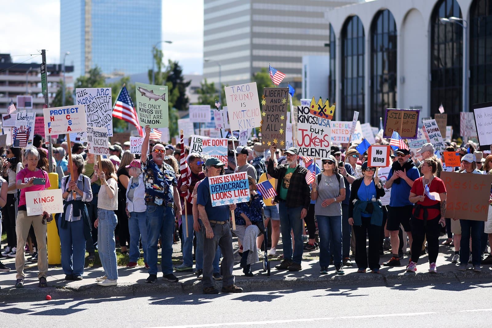Protesters hold signs with political messages during a demonstration in Anchorage, Alaska, on a sunny day in June 2025.