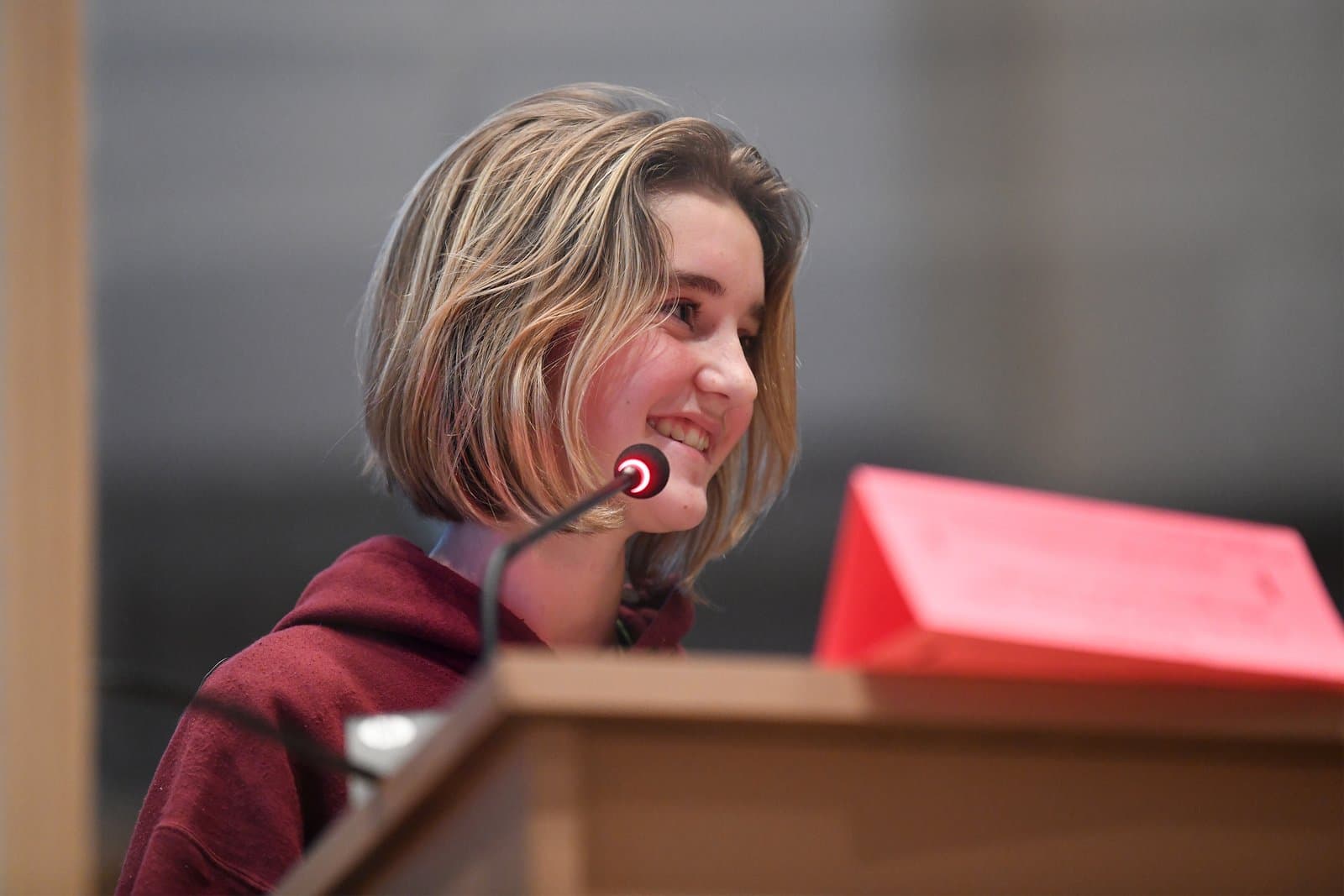 A young person with short blonde hair and a maroon hoodie smiles while speaking into a microphone at a wooden podium during an Anchorage Assembly meeting.